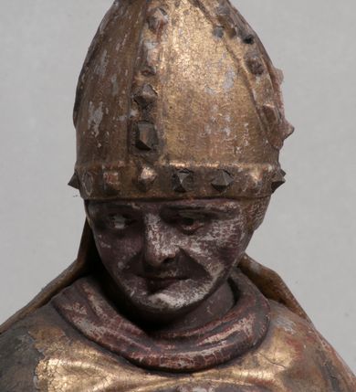 Zdjęcie nr 3: Figura nieopracowana rzeźbiarsko z tyłu, ustawiona jest na niewielkiej, nieregularnej podstawie. Święty ukazany jest w całej postaci, frontalnie, z głową skierowaną w prawo. Prawą rękę unosi delikatnie w geście błogosławieństwa, w lewej wyciągniętej do przodu trzyma laskę zakończoną krzyżem. Jego twarz jest trójkątna, o półprzymkniętych oczach, niewielkim nosie i wąskich ustach. Ubrany jest w albę, ornat z płaskorzeźbioną krzyżową pretekstą, kapę zapiętą na piersi, na głowie ma infułę. Przy jego prawej nodze znajduje się półpostać Piotrowina, zwróconego ku świętemu,  z rękoma złożonymi w geście modlitwy, nakrytego płaszczem. Polichromia w odsłoniętych partiach karnacji obecnie poszarzała, strój złocony. 







