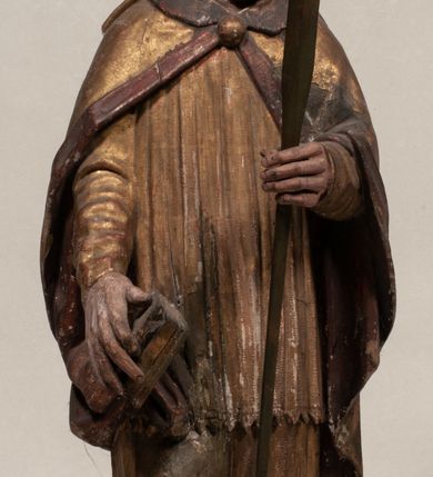 Zdjęcie nr 2: Figura nieopracowana z tyłu, ustawiona na niewielkiej podstawie. Święty ukazany jest w całej postaci, frontalnie, w kontrapoście, z pochyloną głową skierowaną w lewo. W prawej, opuszczonej wzdłuż ciała ręce trzyma zamkniętą księgę, w lewej, uniesionej przed sobą na wysokości piersi – wiosło. Jego twarz jest trójkątna o migdałowatych oczach, krótkim nosie i wąskich ustach, okolona brodą. Ubrany jest w albę, rokietę oraz płaszcz z kołnierzem zapięty pod szyją, na głowie ma infułę. Polichromia naturalistyczna w partiach ciała, strój i atrybuty złocone. 
