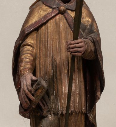 Zdjęcie nr 8: Figura nieopracowana z tyłu, ustawiona na niewielkiej podstawie. Święty ukazany jest w całej postaci, frontalnie, w kontrapoście, z pochyloną głową skierowaną w lewo. W prawej, opuszczonej wzdłuż ciała ręce trzyma zamkniętą księgę, w lewej, uniesionej przed sobą na wysokości piersi – wiosło. Jego twarz jest trójkątna o migdałowatych oczach, krótkim nosie i wąskich ustach, okolona brodą. Ubrany jest w albę, rokietę oraz płaszcz z kołnierzem zapięty pod szyją, na głowie ma infułę. Polichromia naturalistyczna w partiach ciała, strój i atrybuty złocone. 
