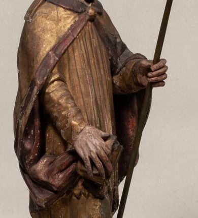 Zdjęcie nr 7: Figura nieopracowana z tyłu, ustawiona na niewielkiej podstawie. Święty ukazany jest w całej postaci, frontalnie, w kontrapoście, z pochyloną głową skierowaną w lewo. W prawej, opuszczonej wzdłuż ciała ręce trzyma zamkniętą księgę, w lewej, uniesionej przed sobą na wysokości piersi – wiosło. Jego twarz jest trójkątna o migdałowatych oczach, krótkim nosie i wąskich ustach, okolona brodą. Ubrany jest w albę, rokietę oraz płaszcz z kołnierzem zapięty pod szyją, na głowie ma infułę. Polichromia naturalistyczna w partiach ciała, strój i atrybuty złocone. 
