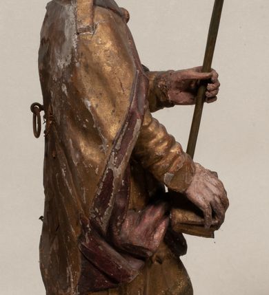 Zdjęcie nr 9: Figura nieopracowana z tyłu, ustawiona na niewielkiej podstawie. Święty ukazany jest w całej postaci, frontalnie, w kontrapoście, z pochyloną głową skierowaną w lewo. W prawej, opuszczonej wzdłuż ciała ręce trzyma zamkniętą księgę, w lewej, uniesionej przed sobą na wysokości piersi – wiosło. Jego twarz jest trójkątna o migdałowatych oczach, krótkim nosie i wąskich ustach, okolona brodą. Ubrany jest w albę, rokietę oraz płaszcz z kołnierzem zapięty pod szyją, na głowie ma infułę. Polichromia naturalistyczna w partiach ciała, strój i atrybuty złocone. 
