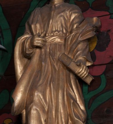 Zdjęcie nr 2: Rzeźba jest ścięta i drążona z tyłu, ustawiona na niskiej podstawie w kształcie półkola. Święty ukazany jest w całej postaci, zwrócony frontalnie, w wykroku. W lewej ręce opuszczonej wzdłuż ciała trzyma zamkniętą księgę, prawa jest ugięta, jej dłoń o zwiniętych palcach jest przyciśnięta do ciała na wysokości pasa. Twarz ma kwadratową, o migdałowatych oczach, krótkim nosie i pełnych ustach, okoloną brodą oraz krótkimi, falowanymi włosami umieszczonymi po bokach głowy, z pasmem włosów nad czołem. Święty ubrany jest w długą tunikę przepasaną szarfą oraz płaszcz narzucony na prawe ramię i lewe przedramię opadający po bokach postaci. Figura w całości polichromowana w kolorze złotym.
