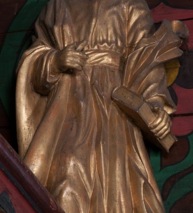 Zdjęcie nr 1: Rzeźba jest ścięta i drążona z tyłu, ustawiona na niskiej podstawie w kształcie półkola. Święty ukazany jest w całej postaci, zwrócony frontalnie, w wykroku. W lewej ręce opuszczonej wzdłuż ciała trzyma zamkniętą księgę, prawa jest ugięta, jej dłoń o zwiniętych palcach jest przyciśnięta do ciała na wysokości pasa. Twarz ma kwadratową, o migdałowatych oczach, krótkim nosie i pełnych ustach, okoloną brodą oraz krótkimi, falowanymi włosami umieszczonymi po bokach głowy, z pasmem włosów nad czołem. Święty ubrany jest w długą tunikę przepasaną szarfą oraz płaszcz narzucony na prawe ramię i lewe przedramię opadający po bokach postaci. Figura w całości polichromowana w kolorze złotym.
