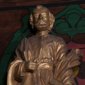 Zdjęcie nr 1: Rzeźba jest ścięta i drążona z tyłu, ustawiona na niskiej podstawie w kształcie półkola. Święty ukazany jest w całej postaci, zwrócony frontalnie, w wykroku. W lewej ręce opuszczonej wzdłuż ciała trzyma zamkniętą księgę, prawa jest ugięta, jej dłoń o zwiniętych palcach jest przyciśnięta do ciała na wysokości pasa. Twarz ma kwadratową, o migdałowatych oczach, krótkim nosie i pełnych ustach, okoloną brodą oraz krótkimi, falowanymi włosami umieszczonymi po bokach głowy, z pasmem włosów nad czołem. Święty ubrany jest w długą tunikę przepasaną szarfą oraz płaszcz narzucony na prawe ramię i lewe przedramię opadający po bokach postaci. Figura w całości polichromowana w kolorze złotym.
