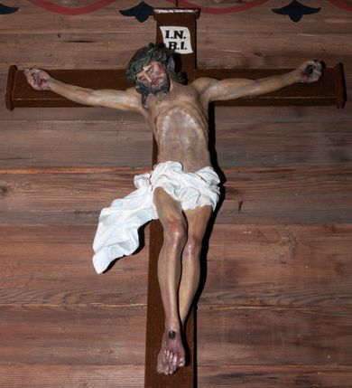 Zdjęcie nr 1: Figura Chrystusa umarłego przybita trzema gwoździami do krzyża o prostych ramionach. Jego ciało jest w delikatnym zwisie o horyzontalnie rozłożonych ramionach. Prawa noga ugięta w kolanie, stopy skrzyżowane w układzie prawa na lewą. Twarz Chrystusa jest szczupła, o głęboko osadzonych, zamkniętych oczach, prostym, krótkim nosie i lekko rozchylonych ustach, okolona krótką brodą skręconą w dwa pukle oraz kręconymi włosami opadającymi na ramiona. Na głowie ma koronę cierniową. Ciało szczupłe, miękko modelowane, o podkreślonej muskulaturze ud i ramion oraz silnie uwypuklonych żebrach i mostku klatki piersiowej. Biodra przesłonięte są białym, krótkim, mocno drapowanym i przylegającym do ciała perizonium z ekspresyjnie rozwianym zwisem tkaniny na prawym boku. Polichromia naturalistyczna z zaznaczonymi śladami męki. Na zakończeniu pionowej belki krzyża namalowana tabliczka z napisem „I.N. / R.I.”.




