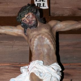 Zdjęcie nr 1: Figura Chrystusa umarłego przybita trzema gwoździami do krzyża o prostych ramionach. Jego ciało jest w delikatnym zwisie o horyzontalnie rozłożonych ramionach. Prawa noga ugięta w kolanie, stopy skrzyżowane w układzie prawa na lewą. Twarz Chrystusa jest szczupła, o głęboko osadzonych, zamkniętych oczach, prostym, krótkim nosie i lekko rozchylonych ustach, okolona krótką brodą skręconą w dwa pukle oraz kręconymi włosami opadającymi na ramiona. Na głowie ma koronę cierniową. Ciało szczupłe, miękko modelowane, o podkreślonej muskulaturze ud i ramion oraz silnie uwypuklonych żebrach i mostku klatki piersiowej. Biodra przesłonięte są białym, krótkim, mocno drapowanym i przylegającym do ciała perizonium z ekspresyjnie rozwianym zwisem tkaniny na prawym boku. Polichromia naturalistyczna z zaznaczonymi śladami męki. Na zakończeniu pionowej belki krzyża namalowana tabliczka z napisem „I.N. / R.I.”.




