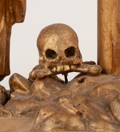 Zdjęcie nr 19: Grupa Ukrzyżowania umieszczona jest na wysokim cokole z ryzalitem pośrodku, profilowanym u dołu i zakończonym gzymsem z kostkowaniem. Do cokołu z trzech stron przytwierdzone są gwoździkami płaskorzeźbione narzędzia Męki Pańskiej. Grupa ustawiona jest na rzeźbionym podłożu imitującym skałę. Pośrodku znajduje się krzyż z pnia drzewa, do którego przybita jest trzema gwoździami figurka umarłego Chrystusa. Jego ciało jest w dużym zwisie, o ramionach przyjmujących kształt łuku półkolistego, głowa opada na prawy bark, nogi ułożone są równolegle, stopy skrzyżowane w układzie prawa na lewą. Twarz pociągła, okolona krótką brodą oraz silnie kręconymi włosami, na głowie korona cierniowa. Ciało smukłe, z podkreślonymi żebrami klatki piersiowej i zaznaczonymi śladami męki. Biodra przepasane krótkim, ekspresyjnie drapowanym perizonium, zawieszonym na podwójnym sznurze, odsłaniającym prawe biodro, zawiązanym na prawym boku. Pionowa belka krzyża zakończona banderolą z rytym napisem „IN / RI”. U podstawy krzyża znajduje się czaszka z piszczelami. 
Po prawej stronie krzyża stoi Maria ze splecionymi z przodu dłońmi. Ubrana jest w tunikę, przepasaną w talii oraz płaszcz narzucony na głowę i ramiona. Po lewej stronie znajduje się św. Jan. Zwrócony jest w trzech czwartych w prawo, klęczy na prawym kolanie i prawą dłoń kładzie na piersi, lewą rękę wyciąga do przodu. Jego twarz okalają długie włosy opadające na ramiona. Ubrany jest w tunikę, przepasaną w talii. 
Figury oraz detale rzeźbiarskie i profile cokołu pozłocone, struktura cokołu polichromowana na czarno. 