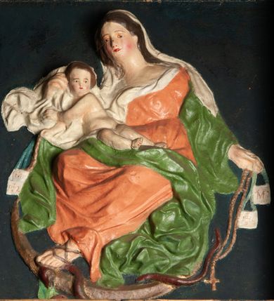 Zdjęcie nr 1: Płaskorzeźba Matki Boskiej z Dzieciątkiem na prostokątnym tle. Maria ukazana jako siedząca, zwrócona w trzech czwartych w prawo, lewą stopę opiera na półksiężycu, depcząc głowę węża.
W lewej, wyciągniętej w bok ręce trzyma szkaplerz i różaniec, prawą przytrzymuje Dzieciątko siedzące na jej kolanie. Twarz ma owalną, o delikatnych rysach, okoloną długimi włosami opadającymi na ramiona. Ubrana jest w pomarańczową suknię o dużym dekolcie, zielony płaszcz zarzucony na przedramiona, opadający po bokach postaci i na kolana, na głowie ma biały welon, z partią tkaniny skumulowaną wokół prawej ręki. Dzieciątko zwrócone jest w trzech czwartych w lewo, z torsem i rękami skierowanymi w przeciwną stronę, opiera się głową o rękę Matki, w prawej rączce trzyma szkaplerz i różaniec. Przedstawienie w prostej ramie zwieńczonej kokardą, ujętej po bokach uszami w formie zębów. Polichromia naturalistyczna. 
