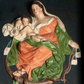 Zdjęcie nr 1: Płaskorzeźba Matki Boskiej z Dzieciątkiem na prostokątnym tle. Maria ukazana jako siedząca, zwrócona w trzech czwartych w prawo, lewą stopę opiera na półksiężycu, depcząc głowę węża.
W lewej, wyciągniętej w bok ręce trzyma szkaplerz i różaniec, prawą przytrzymuje Dzieciątko siedzące na jej kolanie. Twarz ma owalną, o delikatnych rysach, okoloną długimi włosami opadającymi na ramiona. Ubrana jest w pomarańczową suknię o dużym dekolcie, zielony płaszcz zarzucony na przedramiona, opadający po bokach postaci i na kolana, na głowie ma biały welon, z partią tkaniny skumulowaną wokół prawej ręki. Dzieciątko zwrócone jest w trzech czwartych w lewo, z torsem i rękami skierowanymi w przeciwną stronę, opiera się głową o rękę Matki, w prawej rączce trzyma szkaplerz i różaniec. Przedstawienie w prostej ramie zwieńczonej kokardą, ujętej po bokach uszami w formie zębów. Polichromia naturalistyczna. 
