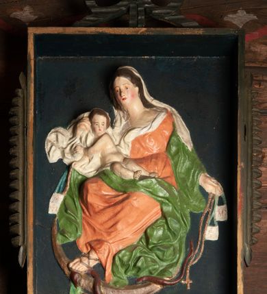 Zdjęcie nr 2: Płaskorzeźba Matki Boskiej z Dzieciątkiem na prostokątnym tle. Maria ukazana jako siedząca, zwrócona w trzech czwartych w prawo, lewą stopę opiera na półksiężycu, depcząc głowę węża.
W lewej, wyciągniętej w bok ręce trzyma szkaplerz i różaniec, prawą przytrzymuje Dzieciątko siedzące na jej kolanie. Twarz ma owalną, o delikatnych rysach, okoloną długimi włosami opadającymi na ramiona. Ubrana jest w pomarańczową suknię o dużym dekolcie, zielony płaszcz zarzucony na przedramiona, opadający po bokach postaci i na kolana, na głowie ma biały welon, z partią tkaniny skumulowaną wokół prawej ręki. Dzieciątko zwrócone jest w trzech czwartych w lewo, z torsem i rękami skierowanymi w przeciwną stronę, opiera się głową o rękę Matki, w prawej rączce trzyma szkaplerz i różaniec. Przedstawienie w prostej ramie zwieńczonej kokardą, ujętej po bokach uszami w formie zębów. Polichromia naturalistyczna. 
