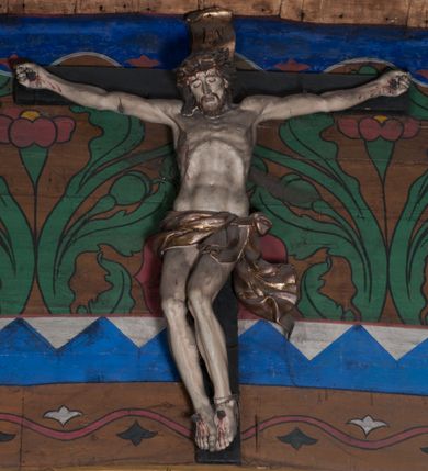 Zdjęcie nr 1: Figura umarłego Chrystusa przybita jest do krzyża o prostych ramionach czterema gwoździami. Ciało w lekkim zwisie o horyzontalnie rozłożonych ramionach, głowa bezwładnie opada na prawe ramię, nogi ugięte w kolanach, skierowane w prawo. Twarz Chrystusa jest pociągła, o zamkniętych oczach, prostym, długim nosie oraz pełnych, rozchylonych ustach, okolona krótką brodą oraz delikatnie kręconymi włosami opadającymi na prawe ramię. Na głowie ma koronę cierniową. Ciało wychudzone, o silnie zaznaczonej muskulaturze rąk i nóg, wyraźnie podkreślonych żebrach klatki piersiowej i zapadłym brzuchu. Biodra przepasane są krótkim perizonium zawieszonym na sznurze, z rozwianym zwisem tkaniny przy lewym boku. Polichromia ciała naturalistyczna z zaznaczonymi śladami męki, korona cierniowa i perizonium pozłocone. 

