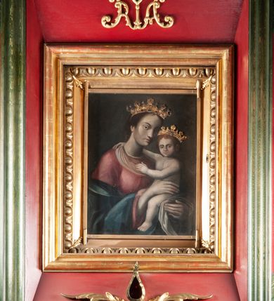 Zdjęcie nr 2: Obraz w kształcie stojącego prostokąta z wizerunkiem Matki Boskiej z Dzieciątkiem. W centrum kompozycji ukazana Maria w półpostaci, delikatnie zwrócona w lewo, z Dzieciątkiem Jezus na lewym ręku, z głową przechyloną w stronę Dzieciątka. Twarz okolona długimi włosami, splecionymi w warkocz z tyłu głowy, nos długi i prosty, drobne usta, wzrok skierowany na widza. Maria ma charakterystyczną długą szyję i smukłe dłonie. Ubrana jest w długą, czerwoną suknię oraz zielony płaszcz, zarzucony na ręce. Dzieciątko ukazane jest w pozycji siedzącej na ramieniu Marii, zwrócone trzy czwarte w prawo, rączkami obejmuje matkę za szyję. Twarz okrągła, nos długi i szeroki, wzrok skierowany na widza, włosy jasne i kędzierzawe. Dzieciątko nagie, siedzi na białej pieluszce. Głowy Marii i Dzieciątka zdobią metalowe, kameryzowane korony w typie otwartym. Tło jednolite, ciemnobrązowe. Rama drewniana, profilowana, złocona, od zewnątrz zdobiona ornamentem w postaci kimationu. Według dokumentacji konserwatorskiej w prawym, dolnym narożu obrazu znajduje się sygnatura „A. Dąbski [lub Dąbrowski] 1678”.
