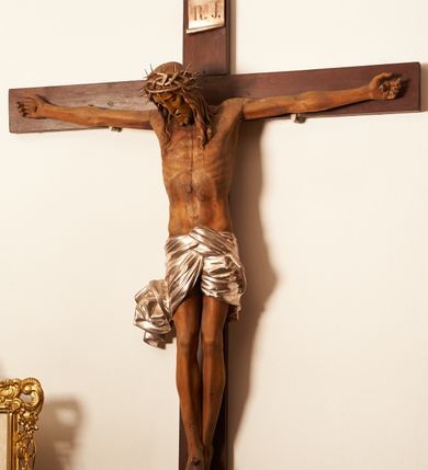 Zdjęcie nr 1: Rzeźbiona figura Chrystusa Ukrzyżowanego zawieszona na prostym, późniejszym od rzeźby krzyżu. Chrustu wisi z rozłożonymi do boków rękami przybitymi gwoździami, ze stopami złożonymi razem, prawa na lewej, przybitymi jednym gwoździem; kolana lekko ugięte. Ciało wyprężone, z zaznaczonymi żebrami i muskulaturą. Głowa opada na prawe ramię, twarz pociągła, oczy przymknięte, proste, nos prosty, długi, usta rozchylone, otoczone krótkimi wąsami i dzieląca się w dwa pukle brodą. Długie, kręcone włosy opadają do przodu, na tors, na głowie masywna, złocona korona cierniowa. Figura bejcowana na kolor ciemnobrązowy. Biodra przewiązane srebrzonym, krótkim perizonium którego zwoje przeplatają się z przodu, a koniec przy prawym udzie zawija się w ekspresyjny zwój. Tkanina układa się w drobne fałdy. Chrystus wisi na drewnianym, ciemnobrązowym krzyżu, w jej górnej części titulus z napisem „I.N./R.I.”.  