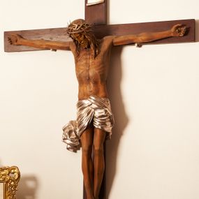 Zdjęcie nr 1: Rzeźbiona figura Chrystusa Ukrzyżowanego zawieszona na prostym, późniejszym od rzeźby krzyżu. Chrustu wisi z rozłożonymi do boków rękami przybitymi gwoździami, ze stopami złożonymi razem, prawa na lewej, przybitymi jednym gwoździem; kolana lekko ugięte. Ciało wyprężone, z zaznaczonymi żebrami i muskulaturą. Głowa opada na prawe ramię, twarz pociągła, oczy przymknięte, proste, nos prosty, długi, usta rozchylone, otoczone krótkimi wąsami i dzieląca się w dwa pukle brodą. Długie, kręcone włosy opadają do przodu, na tors, na głowie masywna, złocona korona cierniowa. Figura bejcowana na kolor ciemnobrązowy. Biodra przewiązane srebrzonym, krótkim perizonium którego zwoje przeplatają się z przodu, a koniec przy prawym udzie zawija się w ekspresyjny zwój. Tkanina układa się w drobne fałdy. Chrystus wisi na drewnianym, ciemnobrązowym krzyżu, w jej górnej części titulus z napisem „I.N./R.I.”.  