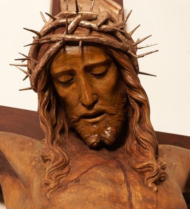 Zdjęcie nr 7: Rzeźbiona figura Chrystusa Ukrzyżowanego zawieszona na prostym, późniejszym od rzeźby krzyżu. Chrustu wisi z rozłożonymi do boków rękami przybitymi gwoździami, ze stopami złożonymi razem, prawa na lewej, przybitymi jednym gwoździem; kolana lekko ugięte. Ciało wyprężone, z zaznaczonymi żebrami i muskulaturą. Głowa opada na prawe ramię, twarz pociągła, oczy przymknięte, proste, nos prosty, długi, usta rozchylone, otoczone krótkimi wąsami i dzieląca się w dwa pukle brodą. Długie, kręcone włosy opadają do przodu, na tors, na głowie masywna, złocona korona cierniowa. Figura bejcowana na kolor ciemnobrązowy. Biodra przewiązane srebrzonym, krótkim perizonium którego zwoje przeplatają się z przodu, a koniec przy prawym udzie zawija się w ekspresyjny zwój. Tkanina układa się w drobne fałdy. Chrystus wisi na drewnianym, ciemnobrązowym krzyżu, w jej górnej części titulus z napisem „I.N./R.I.”.  