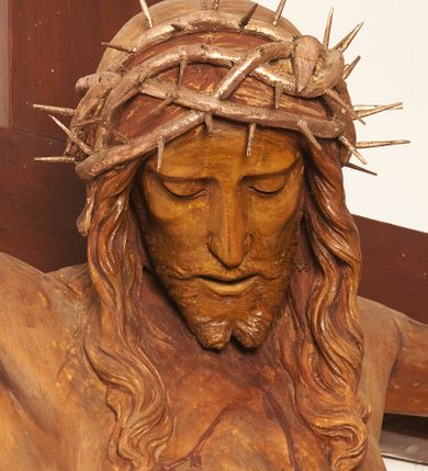 Zdjęcie nr 12: Rzeźbiona figura Chrystusa Ukrzyżowanego zawieszona na prostym, późniejszym od rzeźby krzyżu. Chrustu wisi z rozłożonymi do boków rękami przybitymi gwoździami, ze stopami złożonymi razem, prawa na lewej, przybitymi jednym gwoździem; kolana lekko ugięte. Ciało wyprężone, z zaznaczonymi żebrami i muskulaturą. Głowa opada na prawe ramię, twarz pociągła, oczy przymknięte, proste, nos prosty, długi, usta rozchylone, otoczone krótkimi wąsami i dzieląca się w dwa pukle brodą. Długie, kręcone włosy opadają do przodu, na tors, na głowie masywna, złocona korona cierniowa. Figura bejcowana na kolor ciemnobrązowy. Biodra przewiązane srebrzonym, krótkim perizonium którego zwoje przeplatają się z przodu, a koniec przy prawym udzie zawija się w ekspresyjny zwój. Tkanina układa się w drobne fałdy. Chrystus wisi na drewnianym, ciemnobrązowym krzyżu, w jej górnej części titulus z napisem „I.N./R.I.”.  