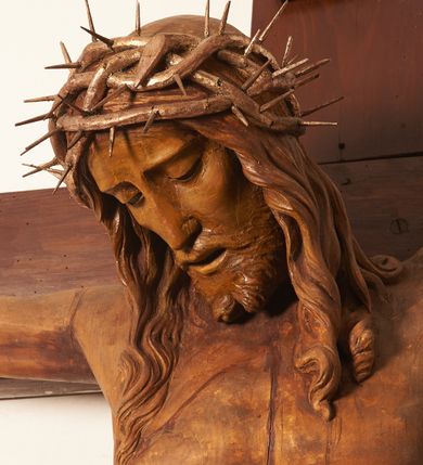 Zdjęcie nr 13: Rzeźbiona figura Chrystusa Ukrzyżowanego zawieszona na prostym, późniejszym od rzeźby krzyżu. Chrustu wisi z rozłożonymi do boków rękami przybitymi gwoździami, ze stopami złożonymi razem, prawa na lewej, przybitymi jednym gwoździem; kolana lekko ugięte. Ciało wyprężone, z zaznaczonymi żebrami i muskulaturą. Głowa opada na prawe ramię, twarz pociągła, oczy przymknięte, proste, nos prosty, długi, usta rozchylone, otoczone krótkimi wąsami i dzieląca się w dwa pukle brodą. Długie, kręcone włosy opadają do przodu, na tors, na głowie masywna, złocona korona cierniowa. Figura bejcowana na kolor ciemnobrązowy. Biodra przewiązane srebrzonym, krótkim perizonium którego zwoje przeplatają się z przodu, a koniec przy prawym udzie zawija się w ekspresyjny zwój. Tkanina układa się w drobne fałdy. Chrystus wisi na drewnianym, ciemnobrązowym krzyżu, w jej górnej części titulus z napisem „I.N./R.I.”.  