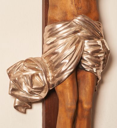 Zdjęcie nr 3: Rzeźbiona figura Chrystusa Ukrzyżowanego zawieszona na prostym, późniejszym od rzeźby krzyżu. Chrustu wisi z rozłożonymi do boków rękami przybitymi gwoździami, ze stopami złożonymi razem, prawa na lewej, przybitymi jednym gwoździem; kolana lekko ugięte. Ciało wyprężone, z zaznaczonymi żebrami i muskulaturą. Głowa opada na prawe ramię, twarz pociągła, oczy przymknięte, proste, nos prosty, długi, usta rozchylone, otoczone krótkimi wąsami i dzieląca się w dwa pukle brodą. Długie, kręcone włosy opadają do przodu, na tors, na głowie masywna, złocona korona cierniowa. Figura bejcowana na kolor ciemnobrązowy. Biodra przewiązane srebrzonym, krótkim perizonium którego zwoje przeplatają się z przodu, a koniec przy prawym udzie zawija się w ekspresyjny zwój. Tkanina układa się w drobne fałdy. Chrystus wisi na drewnianym, ciemnobrązowym krzyżu, w jej górnej części titulus z napisem „I.N./R.I.”.  