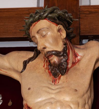 Zdjęcie nr 7: Rzeźba przedstawia Chrystusa Ukrzyżowanego. Postać ukazana jest frontalnie, przybita do krzyża trzema gwoździami z nogami delikatnie ugiętymi w kolanach. Głowa Chrystusa opada na prawe ramię. Sylwetka muskularna z wyraźnie podkreśloną linią żeber i mostka w formie guzów. Twarz podłużna z zamkniętymi oczami, długim i prostym nosem, okolona ciemnobrązową brodą, zwiniętą na końcu w dwa symetryczne pukle. Długie, brązowe włosy opadają na plecy i jednym puklem na prawe ramię. Na głowie Chrystus ma założoną zieloną koronę cierniową. Na prawym boku widać krwawiącą ranę. Podobnie smużki krwi wypływają z ran po gwoździach. Perizonium złocone, przewiązane na prawym boku sznurem i zwisające wzdłuż prawego biodra. W górnej części krzyża znajduje się titulus w formie pionowej banderoli z napisem „IN/RI”. Polichromia w odsłoniętych partiach ciała naturalistyczna. Perizonium i titulus złocone. Krzyż prosty i gładki, malowany na brązowo.

