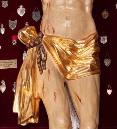 Zdjęcie nr 5: Rzeźba przedstawia Chrystusa Ukrzyżowanego. Postać ukazana jest frontalnie, przybita do krzyża trzema gwoździami z nogami delikatnie ugiętymi w kolanach. Głowa Chrystusa opada na prawe ramię. Sylwetka muskularna z wyraźnie podkreśloną linią żeber i mostka w formie guzów. Twarz podłużna z zamkniętymi oczami, długim i prostym nosem, okolona ciemnobrązową brodą, zwiniętą na końcu w dwa symetryczne pukle. Długie, brązowe włosy opadają na plecy i jednym puklem na prawe ramię. Na głowie Chrystus ma założoną zieloną koronę cierniową. Na prawym boku widać krwawiącą ranę. Podobnie smużki krwi wypływają z ran po gwoździach. Perizonium złocone, przewiązane na prawym boku sznurem i zwisające wzdłuż prawego biodra. W górnej części krzyża znajduje się titulus w formie pionowej banderoli z napisem „IN/RI”. Polichromia w odsłoniętych partiach ciała naturalistyczna. Perizonium i titulus złocone. Krzyż prosty i gładki, malowany na brązowo.

