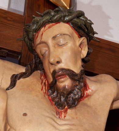 Zdjęcie nr 6: Rzeźba przedstawia Chrystusa Ukrzyżowanego. Postać ukazana jest frontalnie, przybita do krzyża trzema gwoździami z nogami delikatnie ugiętymi w kolanach. Głowa Chrystusa opada na prawe ramię. Sylwetka muskularna z wyraźnie podkreśloną linią żeber i mostka w formie guzów. Twarz podłużna z zamkniętymi oczami, długim i prostym nosem, okolona ciemnobrązową brodą, zwiniętą na końcu w dwa symetryczne pukle. Długie, brązowe włosy opadają na plecy i jednym puklem na prawe ramię. Na głowie Chrystus ma założoną zieloną koronę cierniową. Na prawym boku widać krwawiącą ranę. Podobnie smużki krwi wypływają z ran po gwoździach. Perizonium złocone, przewiązane na prawym boku sznurem i zwisające wzdłuż prawego biodra. W górnej części krzyża znajduje się titulus w formie pionowej banderoli z napisem „IN/RI”. Polichromia w odsłoniętych partiach ciała naturalistyczna. Perizonium i titulus złocone. Krzyż prosty i gładki, malowany na brązowo.

