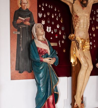 Zdjęcie nr 13: Rzeźba przedstawia Chrystusa Ukrzyżowanego. Postać ukazana jest frontalnie, przybita do krzyża trzema gwoździami z nogami delikatnie ugiętymi w kolanach. Głowa Chrystusa opada na prawe ramię. Sylwetka muskularna z wyraźnie podkreśloną linią żeber i mostka w formie guzów. Twarz podłużna z zamkniętymi oczami, długim i prostym nosem, okolona ciemnobrązową brodą, zwiniętą na końcu w dwa symetryczne pukle. Długie, brązowe włosy opadają na plecy i jednym puklem na prawe ramię. Na głowie Chrystus ma założoną zieloną koronę cierniową. Na prawym boku widać krwawiącą ranę. Podobnie smużki krwi wypływają z ran po gwoździach. Perizonium złocone, przewiązane na prawym boku sznurem i zwisające wzdłuż prawego biodra. W górnej części krzyża znajduje się titulus w formie pionowej banderoli z napisem „IN/RI”. Polichromia w odsłoniętych partiach ciała naturalistyczna. Perizonium i titulus złocone. Krzyż prosty i gładki, malowany na brązowo.

