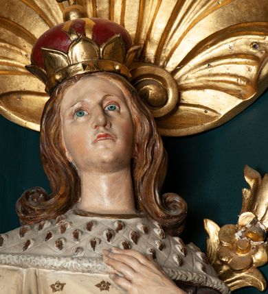 Zdjęcie nr 4: Rzeźba pełnoplastyczna przedstawia św. Kazimierza Królewicza. Święty ukazany jest w pozycji stojącej na niskim cokole, frontalnie, z lewą nogą wysuniętą do przodu. W prawej dłoni trzyma złote berło, lewą ma złożoną na piersi. Twarz podłużna o młodzieńczych rysach, z wyraźnie zaznaczonym podbródkiem, wzrokiem skierowanym przed siebie, okolona długimi i jasnymi włosami, spływającymi równolegle po bokach i symetrycznie podwiniętymi na końcach. Ubrany jest w strój książęcy: długą i białą suknię, obszytą futrem sobolowym, dekorowaną złotymi gwiazdami oraz ciemnoczerwony płaszcz zdobiony wicią akantową z zieloną podszewką, przerzucony swobodnie przez lewe ramię. Na głowie mitra książęca, na stopach buty z wysokimi cholewami, u lewego boku zawieszony na pasie miecz. Polichromia w odsłoniętych partiach ciała naturalistyczna, jasna; berło i detale złocone.

