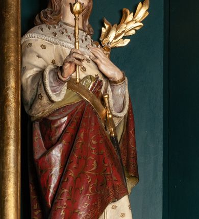 Zdjęcie nr 5: Rzeźba pełnoplastyczna przedstawia św. Kazimierza Królewicza. Święty ukazany jest w pozycji stojącej na niskim cokole, frontalnie, z lewą nogą wysuniętą do przodu. W prawej dłoni trzyma złote berło, lewą ma złożoną na piersi. Twarz podłużna o młodzieńczych rysach, z wyraźnie zaznaczonym podbródkiem, wzrokiem skierowanym przed siebie, okolona długimi i jasnymi włosami, spływającymi równolegle po bokach i symetrycznie podwiniętymi na końcach. Ubrany jest w strój książęcy: długą i białą suknię, obszytą futrem sobolowym, dekorowaną złotymi gwiazdami oraz ciemnoczerwony płaszcz zdobiony wicią akantową z zieloną podszewką, przerzucony swobodnie przez lewe ramię. Na głowie mitra książęca, na stopach buty z wysokimi cholewami, u lewego boku zawieszony na pasie miecz. Polichromia w odsłoniętych partiach ciała naturalistyczna, jasna; berło i detale złocone.

