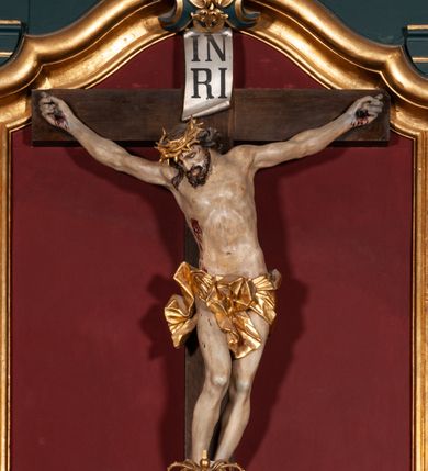 Zdjęcie nr 1: Rzeźba przedstawia Chrystusa Ukrzyżowanego. Postać ukazana jest frontalnie o esowato wygiętej sylwetce, przybita do krzyża trzema gwoździami z nogami ugiętymi w kolanach. Głowa Chrystusa opada na prawe ramię. Sylwetka muskularna z wyraźnie zaznaczoną linią żeber i mostka. Twarz podłużna z niskim czołem, zamkniętymi oczami, długim i prostym nosem oraz wąskimi i lekko rozchylonymi ustami; okolona bujną brodą, zwiniętą na końcu w dwa symetryczne pukle. Długie, brązowe włosy opadają na plecy i prawe ramię, na głowie złota korona cierniowa. Na prawym boku — krwawiąca rana. Podobnie smużki krwi wypływają z ran po gwoździach. Perizonium złocone, silnie drapowane o mocno łamanych fałdach. W górnej części krzyża titulus w formie pionowej banderoli z napisem „IN/RI”. Polichromia w odsłoniętych partiach ciała naturalistyczna. Korona i perizonium złocone, titulus srebrzony. Krzyż prosty i gładki, malowany na brązowo.
