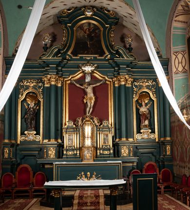 Zdjęcie nr 9: Rzeźba przedstawia Chrystusa Ukrzyżowanego. Postać ukazana jest frontalnie o esowato wygiętej sylwetce, przybita do krzyża trzema gwoździami z nogami ugiętymi w kolanach. Głowa Chrystusa opada na prawe ramię. Sylwetka muskularna z wyraźnie zaznaczoną linią żeber i mostka. Twarz podłużna z niskim czołem, zamkniętymi oczami, długim i prostym nosem oraz wąskimi i lekko rozchylonymi ustami; okolona bujną brodą, zwiniętą na końcu w dwa symetryczne pukle. Długie, brązowe włosy opadają na plecy i prawe ramię, na głowie złota korona cierniowa. Na prawym boku — krwawiąca rana. Podobnie smużki krwi wypływają z ran po gwoździach. Perizonium złocone, silnie drapowane o mocno łamanych fałdach. W górnej części krzyża titulus w formie pionowej banderoli z napisem „IN/RI”. Polichromia w odsłoniętych partiach ciała naturalistyczna. Korona i perizonium złocone, titulus srebrzony. Krzyż prosty i gładki, malowany na brązowo.
