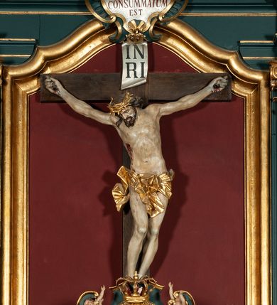 Zdjęcie nr 2: Rzeźba przedstawia Chrystusa Ukrzyżowanego. Postać ukazana jest frontalnie o esowato wygiętej sylwetce, przybita do krzyża trzema gwoździami z nogami ugiętymi w kolanach. Głowa Chrystusa opada na prawe ramię. Sylwetka muskularna z wyraźnie zaznaczoną linią żeber i mostka. Twarz podłużna z niskim czołem, zamkniętymi oczami, długim i prostym nosem oraz wąskimi i lekko rozchylonymi ustami; okolona bujną brodą, zwiniętą na końcu w dwa symetryczne pukle. Długie, brązowe włosy opadają na plecy i prawe ramię, na głowie złota korona cierniowa. Na prawym boku — krwawiąca rana. Podobnie smużki krwi wypływają z ran po gwoździach. Perizonium złocone, silnie drapowane o mocno łamanych fałdach. W górnej części krzyża titulus w formie pionowej banderoli z napisem „IN/RI”. Polichromia w odsłoniętych partiach ciała naturalistyczna. Korona i perizonium złocone, titulus srebrzony. Krzyż prosty i gładki, malowany na brązowo.
