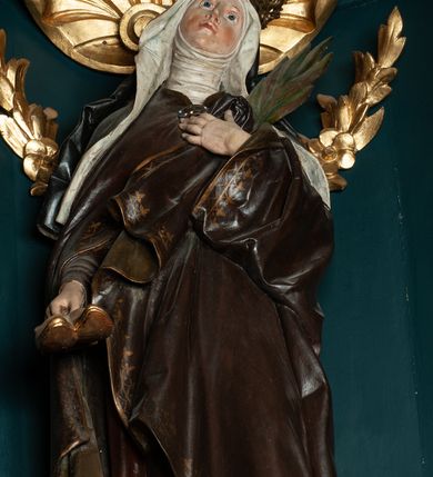 Zdjęcie nr 5: Rzeźba pełnoplastyczna przedstawiająca św. Jadwigę Śląską. Święta ukazana jest w pozycji stojącej na niskim cokole, frontalnie, w delikatnym kontrapoście z lewą nogą ugiętą w kolanie, z głową uniesioną do góry i nieznacznie skierowaną w swoją lewą stronę. W prawej dłoni trzyma parę butów; lewą dłonią, którą ma złożoną na piersi przyciska palmę męczeństwa. Twarz owalna, o spokojnym wyrazie, nos długi i wąski, wzrok wpatrzony w górę, delikatnie zarysowane łuki brwiowe, silnie zarumienione policzki. Ubrana w strój zakonny: ciemno-brązowy habit z białym welonem oraz ciemnobrązowy płaszcz spięty na piersi klamrą, zdobioną kamieniami, na głowie złota korona zamknięta. Fałdy habitu, silnie drapowane opadają ciężko w dół. Polichromia w odsłoniętych partiach ciała naturalistyczna, jasna.

