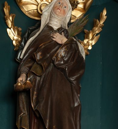 Zdjęcie nr 4: Rzeźba pełnoplastyczna przedstawiająca św. Jadwigę Śląską. Święta ukazana jest w pozycji stojącej na niskim cokole, frontalnie, w delikatnym kontrapoście z lewą nogą ugiętą w kolanie, z głową uniesioną do góry i nieznacznie skierowaną w swoją lewą stronę. W prawej dłoni trzyma parę butów; lewą dłonią, którą ma złożoną na piersi przyciska palmę męczeństwa. Twarz owalna, o spokojnym wyrazie, nos długi i wąski, wzrok wpatrzony w górę, delikatnie zarysowane łuki brwiowe, silnie zarumienione policzki. Ubrana w strój zakonny: ciemno-brązowy habit z białym welonem oraz ciemnobrązowy płaszcz spięty na piersi klamrą, zdobioną kamieniami, na głowie złota korona zamknięta. Fałdy habitu, silnie drapowane opadają ciężko w dół. Polichromia w odsłoniętych partiach ciała naturalistyczna, jasna.

