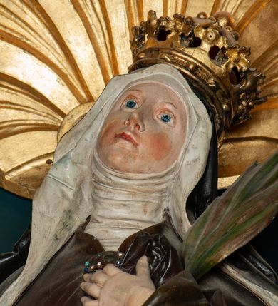 Zdjęcie nr 2: Rzeźba pełnoplastyczna przedstawiająca św. Jadwigę Śląską. Święta ukazana jest w pozycji stojącej na niskim cokole, frontalnie, w delikatnym kontrapoście z lewą nogą ugiętą w kolanie, z głową uniesioną do góry i nieznacznie skierowaną w swoją lewą stronę. W prawej dłoni trzyma parę butów; lewą dłonią, którą ma złożoną na piersi przyciska palmę męczeństwa. Twarz owalna, o spokojnym wyrazie, nos długi i wąski, wzrok wpatrzony w górę, delikatnie zarysowane łuki brwiowe, silnie zarumienione policzki. Ubrana w strój zakonny: ciemno-brązowy habit z białym welonem oraz ciemnobrązowy płaszcz spięty na piersi klamrą, zdobioną kamieniami, na głowie złota korona zamknięta. Fałdy habitu, silnie drapowane opadają ciężko w dół. Polichromia w odsłoniętych partiach ciała naturalistyczna, jasna.

