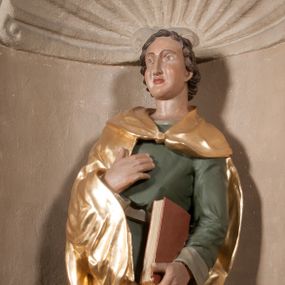 Zdjęcie nr 1: Rzeźba drążona, postać umieszczona na cokole. Święty Jan Ewangelista ukazany w całej postaci, frontalnie, prawą rękę przyciska do piersi w lewej opuszczonej wzdłuż ciała trzyma zamkniętą księgę. Twarz pociągła, o pełnych policzkach z długim nosem, małymi ustami oraz jasnoniebieskimi oczami, okolona krótkimi brązowymi włosami układającymi się w pukle. Święty jest ubrany w zieloną tunikę z wąskimi rękawami opadającą do podstawy płytkimi, miękkimi fałdami oraz złoty płaszcz spięty na dekolcie, zarzucony na ramiona, podtrzymywany z przodu przez prawe przedramię. Obok bosych stóp świętego, po jego prawej stronie ukazany biały orzeł z rozpostartymi skrzydłami.