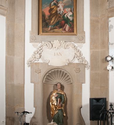 Zdjęcie nr 2: Rzeźba drążona, postać umieszczona na cokole. Święty Jan Ewangelista ukazany w całej postaci, frontalnie, prawą rękę przyciska do piersi w lewej opuszczonej wzdłuż ciała trzyma zamkniętą księgę. Twarz pociągła, o pełnych policzkach z długim nosem, małymi ustami oraz jasnoniebieskimi oczami, okolona krótkimi brązowymi włosami układającymi się w pukle. Święty jest ubrany w zieloną tunikę z wąskimi rękawami opadającą do podstawy płytkimi, miękkimi fałdami oraz złoty płaszcz spięty na dekolcie, zarzucony na ramiona, podtrzymywany z przodu przez prawe przedramię. Obok bosych stóp świętego, po jego prawej stronie ukazany biały orzeł z rozpostartymi skrzydłami.