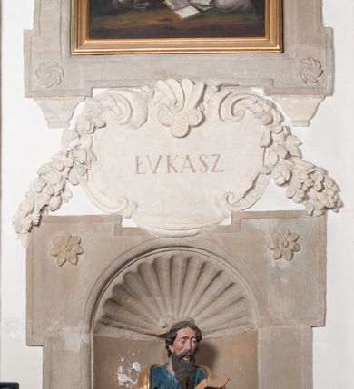 Zdjęcie nr 3: Rzeźba drążona, postać umieszczona na cokole, przedstawiająca św. Łukasza Ewangelistę. Święty ukazany jest w pozycji stojącej, z górną partią ciała skręconą nieznacznie w lewo, z lekko pochyloną głową. Ręce ma ugięte, w lewej trzyma otwartą księgę, prawą wyciąga przed siebie. Ma pociągłą twarz z okrągłymi oczami, długim nosem i wydatnymi ustami, okoloną krótkimi, opadającymi do tyłu puklami włosów oraz długą, dzielącą się na końcu w dwa pukle brodę. Święty odziany jest w przewiązaną w pasie niebieską suknię opadającą do stóp płytkimi, miękkimi fałdami oraz złoty płaszcz, zarzucony na ramiona i otaczający biodra, przytrzymany z przodu rękami, układający się w głębokie, nieregularne fałdy. U bosych stóp świętego wół.