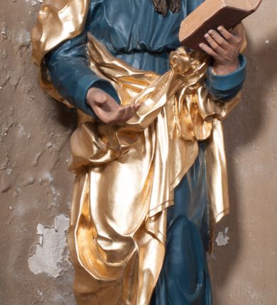Zdjęcie nr 1: Rzeźba drążona, postać umieszczona na cokole, przedstawiająca św. Łukasza Ewangelistę. Święty ukazany jest w pozycji stojącej, z górną partią ciała skręconą nieznacznie w lewo, z lekko pochyloną głową. Ręce ma ugięte, w lewej trzyma otwartą księgę, prawą wyciąga przed siebie. Ma pociągłą twarz z okrągłymi oczami, długim nosem i wydatnymi ustami, okoloną krótkimi, opadającymi do tyłu puklami włosów oraz długą, dzielącą się na końcu w dwa pukle brodę. Święty odziany jest w przewiązaną w pasie niebieską suknię opadającą do stóp płytkimi, miękkimi fałdami oraz złoty płaszcz, zarzucony na ramiona i otaczający biodra, przytrzymany z przodu rękami, układający się w głębokie, nieregularne fałdy. U bosych stóp świętego wół.