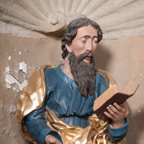 Zdjęcie nr 1: Rzeźba drążona, postać umieszczona na cokole, przedstawiająca św. Łukasza Ewangelistę. Święty ukazany jest w pozycji stojącej, z górną partią ciała skręconą nieznacznie w lewo, z lekko pochyloną głową. Ręce ma ugięte, w lewej trzyma otwartą księgę, prawą wyciąga przed siebie. Ma pociągłą twarz z okrągłymi oczami, długim nosem i wydatnymi ustami, okoloną krótkimi, opadającymi do tyłu puklami włosów oraz długą, dzielącą się na końcu w dwa pukle brodę. Święty odziany jest w przewiązaną w pasie niebieską suknię opadającą do stóp płytkimi, miękkimi fałdami oraz złoty płaszcz, zarzucony na ramiona i otaczający biodra, przytrzymany z przodu rękami, układający się w głębokie, nieregularne fałdy. U bosych stóp świętego wół.