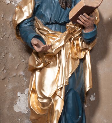 Zdjęcie nr 6: Rzeźba drążona, postać umieszczona na cokole, przedstawiająca św. Łukasza Ewangelistę. Święty ukazany jest w pozycji stojącej, z górną partią ciała skręconą nieznacznie w lewo, z lekko pochyloną głową. Ręce ma ugięte, w lewej trzyma otwartą księgę, prawą wyciąga przed siebie. Ma pociągłą twarz z okrągłymi oczami, długim nosem i wydatnymi ustami, okoloną krótkimi, opadającymi do tyłu puklami włosów oraz długą, dzielącą się na końcu w dwa pukle brodę. Święty odziany jest w przewiązaną w pasie niebieską suknię opadającą do stóp płytkimi, miękkimi fałdami oraz złoty płaszcz, zarzucony na ramiona i otaczający biodra, przytrzymany z przodu rękami, układający się w głębokie, nieregularne fałdy. U bosych stóp świętego wół.