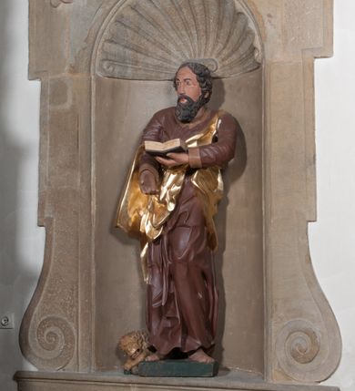 Zdjęcie nr 5: Rzeźba drążona, postać umieszczona na cokole przedstawiająca św. Marka Ewangelistę. Święty ukazany jest w pozycji stojącej, w kontrapoście, z lewą nogą zgiętą w kolanie i sylwetką przechyloną nieznacznie na lewą stronę, a głową odwróconą w przeciwnym kierunku. Prawa ręka wyprostowana, wyciągnięta przed siebie, lewa ugięta, podtrzymuje otwartą księgę. Święty ma pociągłą twarz o okrągłych, szeroko otwartych, niebieskich oczach, długim, wąskim nosie i małych ustach, okoloną zwiniętymi w pukle włosami zakrywającymi uszy oraz od dołu silnie poskręcaną brodę. Ubrany jest w brązową suknię z podwiniętymi rękawami opadającą do stóp podkreślającymi anatomię fałdami oraz w złoty płaszcz przechodzący przez lewy bark i zarzucony na prawe przedramię, układający się w nieregularne, głębokie fałdy. Stopy bose. Przy prawej nodze świętego lew. 
