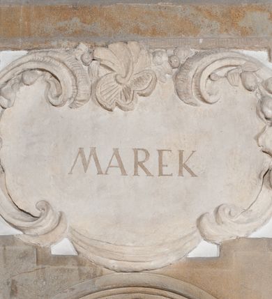 Zdjęcie nr 2: Rzeźba drążona, postać umieszczona na cokole przedstawiająca św. Marka Ewangelistę. Święty ukazany jest w pozycji stojącej, w kontrapoście, z lewą nogą zgiętą w kolanie i sylwetką przechyloną nieznacznie na lewą stronę, a głową odwróconą w przeciwnym kierunku. Prawa ręka wyprostowana, wyciągnięta przed siebie, lewa ugięta, podtrzymuje otwartą księgę. Święty ma pociągłą twarz o okrągłych, szeroko otwartych, niebieskich oczach, długim, wąskim nosie i małych ustach, okoloną zwiniętymi w pukle włosami zakrywającymi uszy oraz od dołu silnie poskręcaną brodę. Ubrany jest w brązową suknię z podwiniętymi rękawami opadającą do stóp podkreślającymi anatomię fałdami oraz w złoty płaszcz przechodzący przez lewy bark i zarzucony na prawe przedramię, układający się w nieregularne, głębokie fałdy. Stopy bose. Przy prawej nodze świętego lew. 