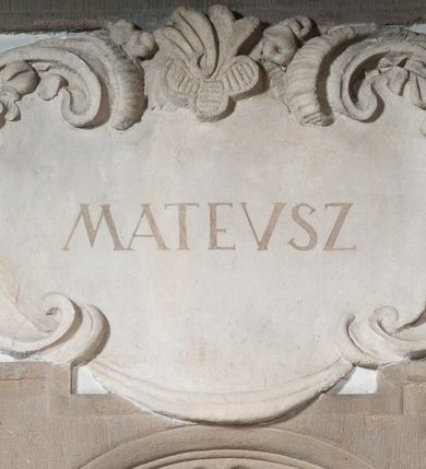 Zdjęcie nr 1: Rzeźba drążona, postać umieszczona na cokole przedstawiająca św. Mateusza. Święty ukazany w pozycji stojącej, w kontrapoście, z lewą nogą odwiedzioną do tyłu i z głową skręconą lekko w lewą stronę. Prawa ręka wyprostowana, sięga w dół w kierunku drugiej postaci (pełniącej rolę atrybutu na dole, wyobrażonej przy nodze świętego), lewa ugięta, podtrzymuje otwartą księgę. Pociągłą twarz świętego okala ciemna, długa, falowana broda i wąsy. Włosy krótkie, kręcone. Święty jest ubrany w beżową suknię z podwiniętymi rękawami, przewiązaną w pasie oraz złoty płaszcz zarzucony na lewy bark i przechodzący przez prawe ramię na lewe biodro, gdzie łamie się on w twarde fałdy. Dwa końce tkaniny spływają kaskadowo w dół. Stopy bose. Przy lewej nodze świętego proporcjonalnie zmniejszona postać ludzka ukazana frontalnie, w pozycji stojącej, z prawą  nogą wysuniętą do przodu i z prawą ręką uniesioną ku górze. Jasne, kręcone włosy okalają okrągłą, pogodną twarz. Postać ubrana w czerwoną suknię sięgającą kolan oraz złoty płaszcz, przerzucony przez lewe ramię.