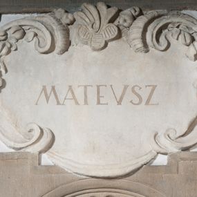 Zdjęcie nr 1: Rzeźba drążona, postać umieszczona na cokole przedstawiająca św. Mateusza. Święty ukazany w pozycji stojącej, w kontrapoście, z lewą nogą odwiedzioną do tyłu i z głową skręconą lekko w lewą stronę. Prawa ręka wyprostowana, sięga w dół w kierunku drugiej postaci (pełniącej rolę atrybutu na dole, wyobrażonej przy nodze świętego), lewa ugięta, podtrzymuje otwartą księgę. Pociągłą twarz świętego okala ciemna, długa, falowana broda i wąsy. Włosy krótkie, kręcone. Święty jest ubrany w beżową suknię z podwiniętymi rękawami, przewiązaną w pasie oraz złoty płaszcz zarzucony na lewy bark i przechodzący przez prawe ramię na lewe biodro, gdzie łamie się on w twarde fałdy. Dwa końce tkaniny spływają kaskadowo w dół. Stopy bose. Przy lewej nodze świętego proporcjonalnie zmniejszona postać ludzka ukazana frontalnie, w pozycji stojącej, z prawą  nogą wysuniętą do przodu i z prawą ręką uniesioną ku górze. Jasne, kręcone włosy okalają okrągłą, pogodną twarz. Postać ubrana w czerwoną suknię sięgającą kolan oraz złoty płaszcz, przerzucony przez lewe ramię.