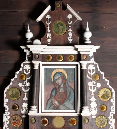 Zdjęcie nr 1: Ołtarzyk architektoniczny, jednokondygnacyjny ze zwieńczeniem, na cokole; w formie aediculi, ujętej parą kolumienek ustawionych na wyodrębnionych cokołach, dźwigających pełne belkowanie wyłamane na ich osi, na którym znajdują się kule i tralki. W polu głównym wnęka w kształcie stojącego prostokąta, ujęta profilowaną ramą z wizerunkiem Matki Boskiej. W partii cokołu pięć medalionów, z których cztery zdobione są rozetami z papieru, środkowa jest pusta. Fryz dekorowany pięcioma medalionami z papierowymi rozetami. Ołtarzyk ujęty skrzydłami o fantazyjnym wykroju, z malowanym wzorem wzdłuż krawędzi. Każde ozdobione czterema medalionami z malowanymi popiersiami apostołów (trzech ewangelistów i św. Macieja) i papierowymi rozetami oraz płaskorzeźbionym ornamentem kandelabrowym. Zwieńczenie o fantazyjnej formie zbliżonej do kwadratu o wklęsłych bokach i ściętych górnych narożach obwiedzionych gzymsem, zakończone małym szczytem z rozetą. W polu medalion z przedstawieniem męczeństwa św. Sebastiana, flankowany płaskorzeźbionym ornamentem kandelabrowym. 
Obraz Matki Boskiej w formie stojącego prostokąta. W centrum półpostać Marii, zwróconej w trzech czwartych w prawo, z pochyloną głową i dłońmi złożonymi w geście modlitwy. Twarz ma owalną, o pełnych policzkach, okrągłych, półprzymkniętych oczach, prostym nosie i małych, pełnych ustach. Ubrana jest w brązową suknię, pod szyją ma podwikę, na ramiona i głowę zarzucony niebieski płaszcz z czerwoną podszewką. Wokół jej głowy złocony nimb. Maria ukazana jest na tle niebiesko-różowego nieba. 











