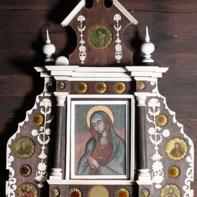 Zdjęcie nr 1: Ołtarzyk architektoniczny, jednokondygnacyjny ze zwieńczeniem, na cokole; w formie aediculi, ujętej parą kolumienek ustawionych na wyodrębnionych cokołach, dźwigających pełne belkowanie wyłamane na ich osi, na którym znajdują się kule i tralki. W polu głównym wnęka w kształcie stojącego prostokąta, ujęta profilowaną ramą z wizerunkiem Matki Boskiej. W partii cokołu pięć medalionów, z których cztery zdobione są rozetami z papieru, środkowa jest pusta. Fryz dekorowany pięcioma medalionami z papierowymi rozetami. Ołtarzyk ujęty skrzydłami o fantazyjnym wykroju, z malowanym wzorem wzdłuż krawędzi. Każde ozdobione czterema medalionami z malowanymi popiersiami apostołów (trzech ewangelistów i św. Macieja) i papierowymi rozetami oraz płaskorzeźbionym ornamentem kandelabrowym. Zwieńczenie o fantazyjnej formie zbliżonej do kwadratu o wklęsłych bokach i ściętych górnych narożach obwiedzionych gzymsem, zakończone małym szczytem z rozetą. W polu medalion z przedstawieniem męczeństwa św. Sebastiana, flankowany płaskorzeźbionym ornamentem kandelabrowym. 
Obraz Matki Boskiej w formie stojącego prostokąta. W centrum półpostać Marii, zwróconej w trzech czwartych w prawo, z pochyloną głową i dłońmi złożonymi w geście modlitwy. Twarz ma owalną, o pełnych policzkach, okrągłych, półprzymkniętych oczach, prostym nosie i małych, pełnych ustach. Ubrana jest w brązową suknię, pod szyją ma podwikę, na ramiona i głowę zarzucony niebieski płaszcz z czerwoną podszewką. Wokół jej głowy złocony nimb. Maria ukazana jest na tle niebiesko-różowego nieba. 











