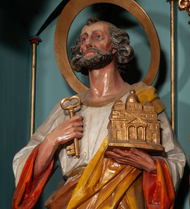 Zdjęcie nr 3: Rzeźba św. Piotra ustawiona na niskim prostopadłościennym cokole, o ściętych narożach. Święty Piotr ukazany frontalnie, w delikatnym kontrapoście, z lewą nogą ugiętą w kolanie. W prawej dłoni trzyma jeden klucz, a w lewej model bazyliki św. Piotra w Rzymie. Twarz podłużna, z długim i szerokim nosem, dużymi oczami i szerokimi ustami, okolona średniej długości, siwą  brodą. Włosy siwe, gęste i zasłaniające uszy, ułożone po bokach w bujne loki. Ubrany jest w długą białą suknię z długimi rękawami, pomarańczową podszewką, przewiązaną w talii oraz żółty płaszcz z brązową podszewką. Wokół głowy ma duży, szeroki, okrągły nimb. Polichromia w odsłoniętych partiach ciała naturalistyczna, detale złocone.