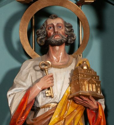 Zdjęcie nr 1: Rzeźba św. Piotra ustawiona na niskim prostopadłościennym cokole, o ściętych narożach. Święty Piotr ukazany frontalnie, w delikatnym kontrapoście, z lewą nogą ugiętą w kolanie. W prawej dłoni trzyma jeden klucz, a w lewej model bazyliki św. Piotra w Rzymie. Twarz podłużna, z długim i szerokim nosem, dużymi oczami i szerokimi ustami, okolona średniej długości, siwą  brodą. Włosy siwe, gęste i zasłaniające uszy, ułożone po bokach w bujne loki. Ubrany jest w długą białą suknię z długimi rękawami, pomarańczową podszewką, przewiązaną w talii oraz żółty płaszcz z brązową podszewką. Wokół głowy ma duży, szeroki, okrągły nimb. Polichromia w odsłoniętych partiach ciała naturalistyczna, detale złocone.