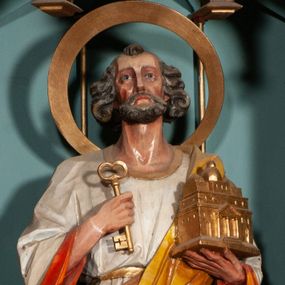 Zdjęcie nr 1: Rzeźba św. Piotra ustawiona na niskim prostopadłościennym cokole, o ściętych narożach. Święty Piotr ukazany frontalnie, w delikatnym kontrapoście, z lewą nogą ugiętą w kolanie. W prawej dłoni trzyma jeden klucz, a w lewej model bazyliki św. Piotra w Rzymie. Twarz podłużna, z długim i szerokim nosem, dużymi oczami i szerokimi ustami, okolona średniej długości, siwą  brodą. Włosy siwe, gęste i zasłaniające uszy, ułożone po bokach w bujne loki. Ubrany jest w długą białą suknię z długimi rękawami, pomarańczową podszewką, przewiązaną w talii oraz żółty płaszcz z brązową podszewką. Wokół głowy ma duży, szeroki, okrągły nimb. Polichromia w odsłoniętych partiach ciała naturalistyczna, detale złocone.