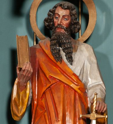 Zdjęcie nr 1: Rzeźba św. Pawła ustawiona na niskim prostopadłościennym cokole o ściętych narożach. Święty ukazany jest frontalnie, w prawej dłoni trzyma księgę, a w lewej miecz skierowany ostrzem do dołu. Twarz o jasnej karnacji, z długim i szerokim nosem, okolona długą i bujną brodą rozdzieloną na dwa pukle, silnie fakturowaną w poziome bruzdy. Włosy ciemnobrązowe, gęste, o bujnych puklach ułożonych po bokach głowy. Ubrany jest w białą suknię z długimi rękawami i kołnierzem oraz jasnobrązowy płaszcz odsłaniający lewe ramię. Wokół głowy ma duży i szeroki, okrągły nimb. Polichromia w odsłoniętych partiach ciała naturalistyczna, detale złocone i srebrzone.
